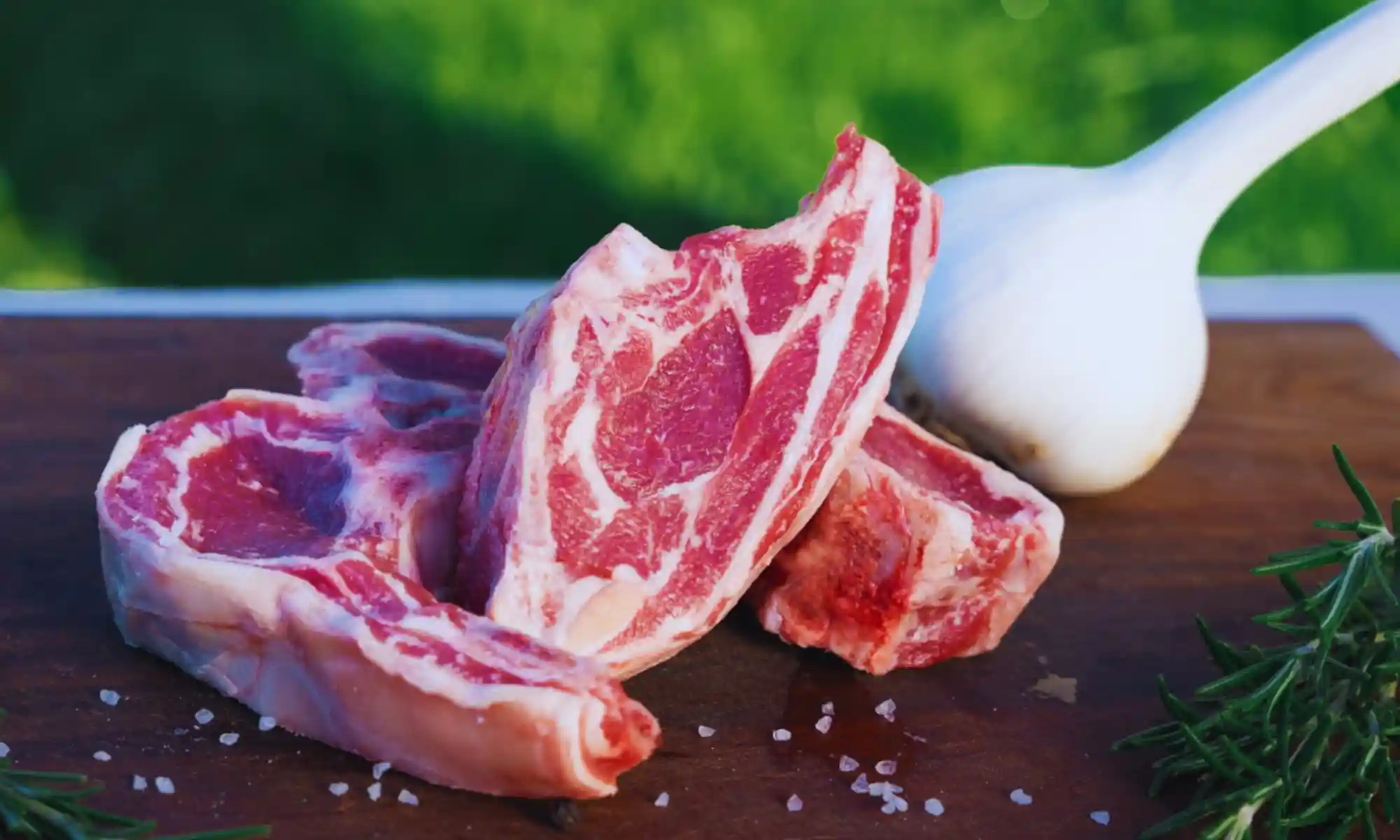 Raw grass-fed lamb chops displayed with garlic and herbs on a wooden board.
